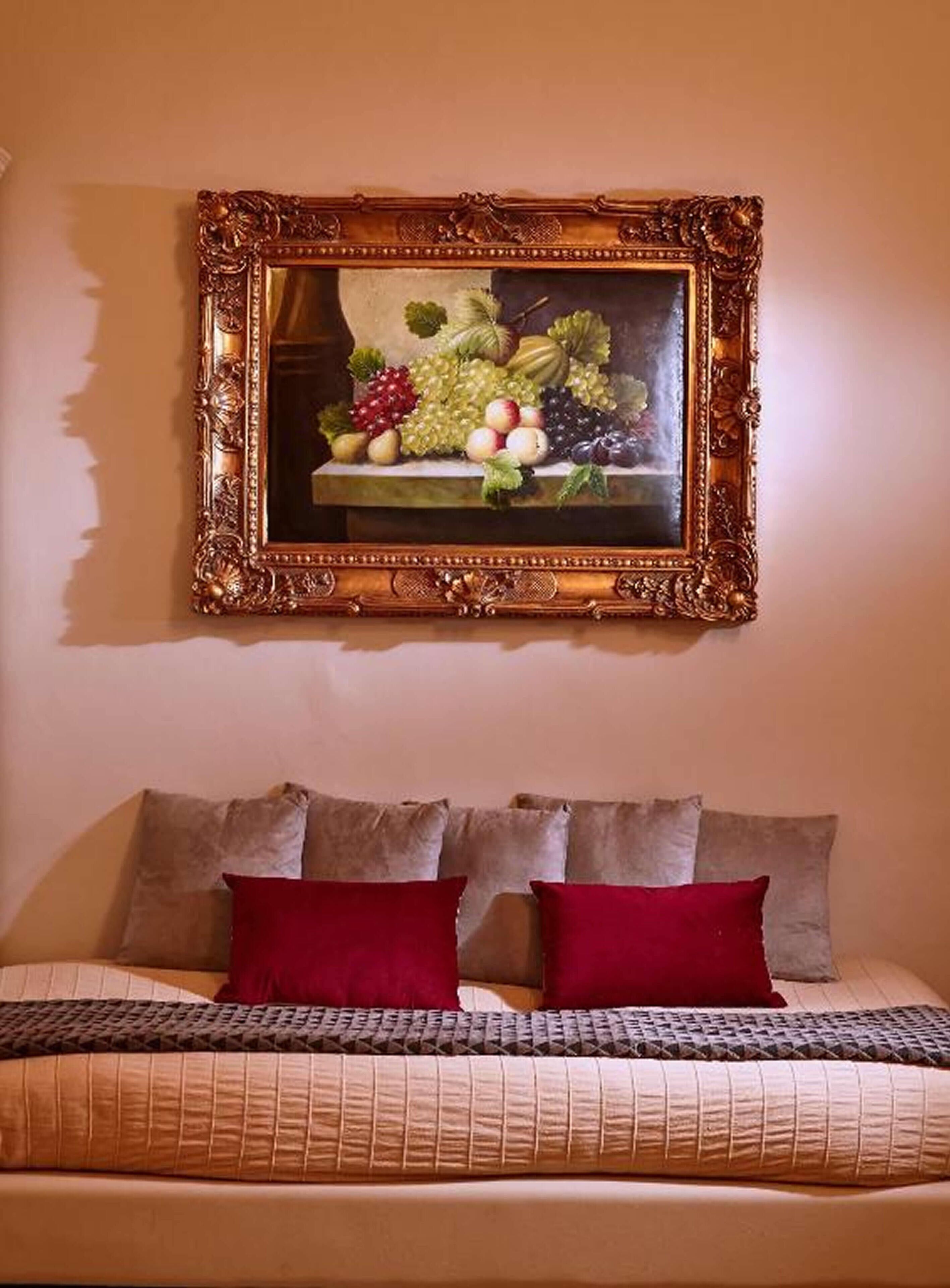 Un lit avec une couvre-lit à motifs et deux oreillers rouges, un tableau représentant des fruits accroché au mur au-dessus, et un mur uni.