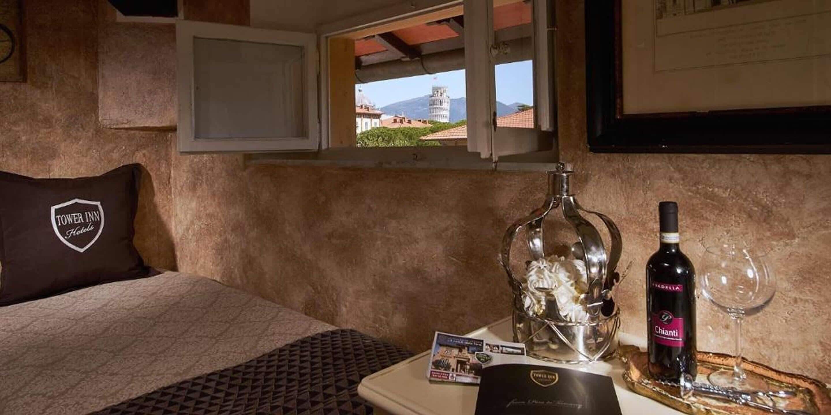 A hotel room with a bed, a window with a view, a bottle of Chianti wine, and a glass.