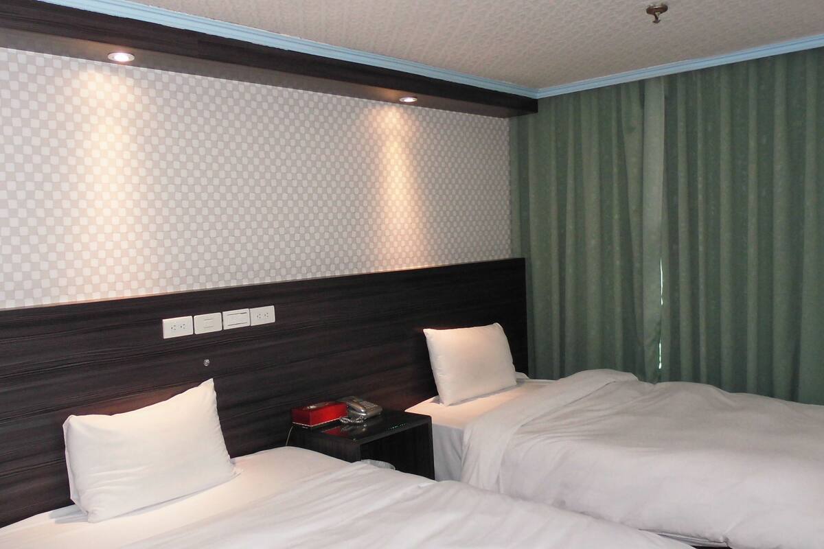 Habitación de hotel con dos camas, cabecera oscura, pared estampada y cortinas verdes.