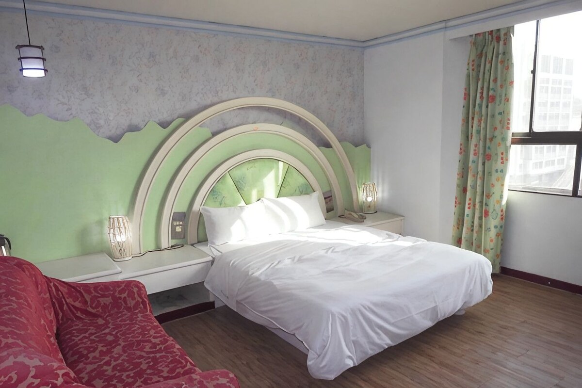 A hotel room with a bed, a bedside table, a red sofa, and a window with curtains.