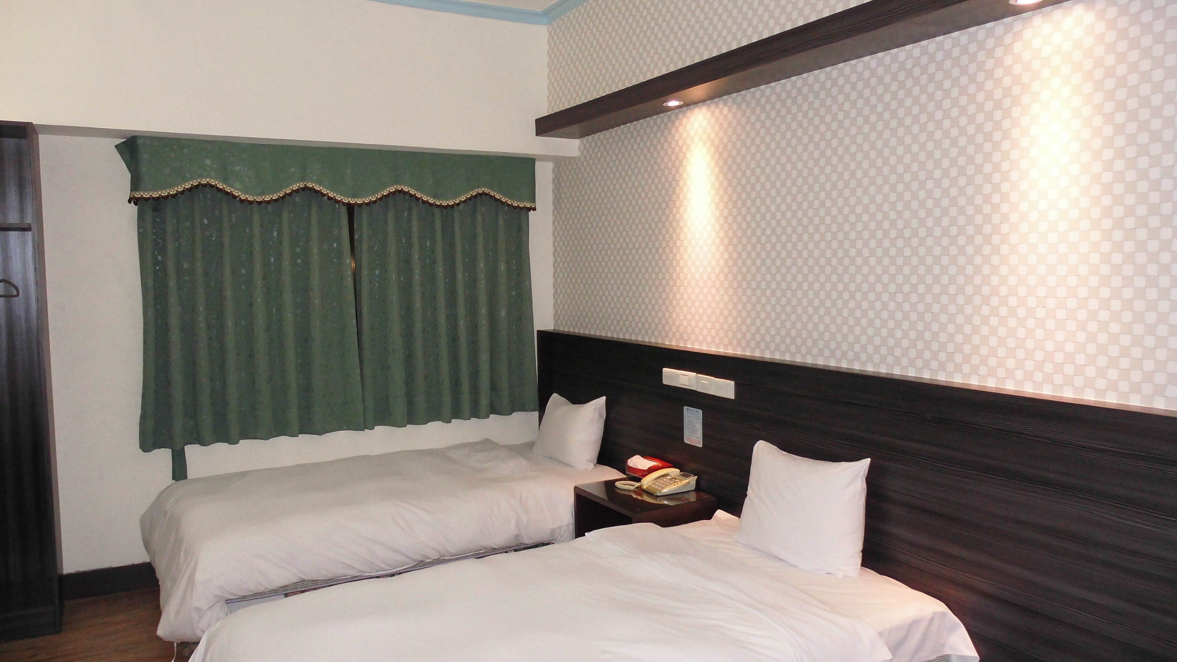 A hotel room with two beds, a green curtain, a patterned wall, and a bedside table with a phone.
