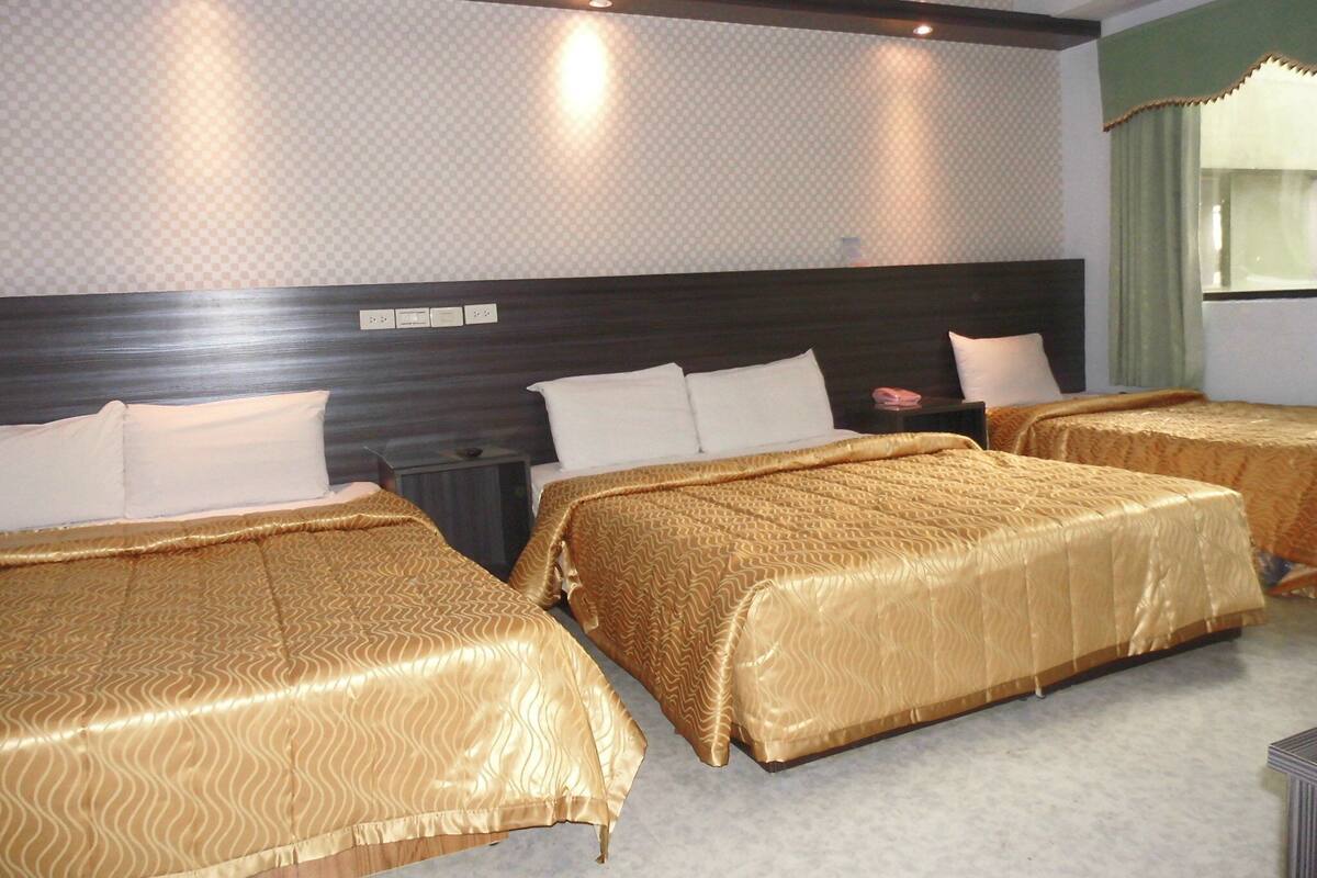 Habitación de hotel con tres camas individuales, cabecera oscura, mesita de noche pequeña y una ventana con cortinas.