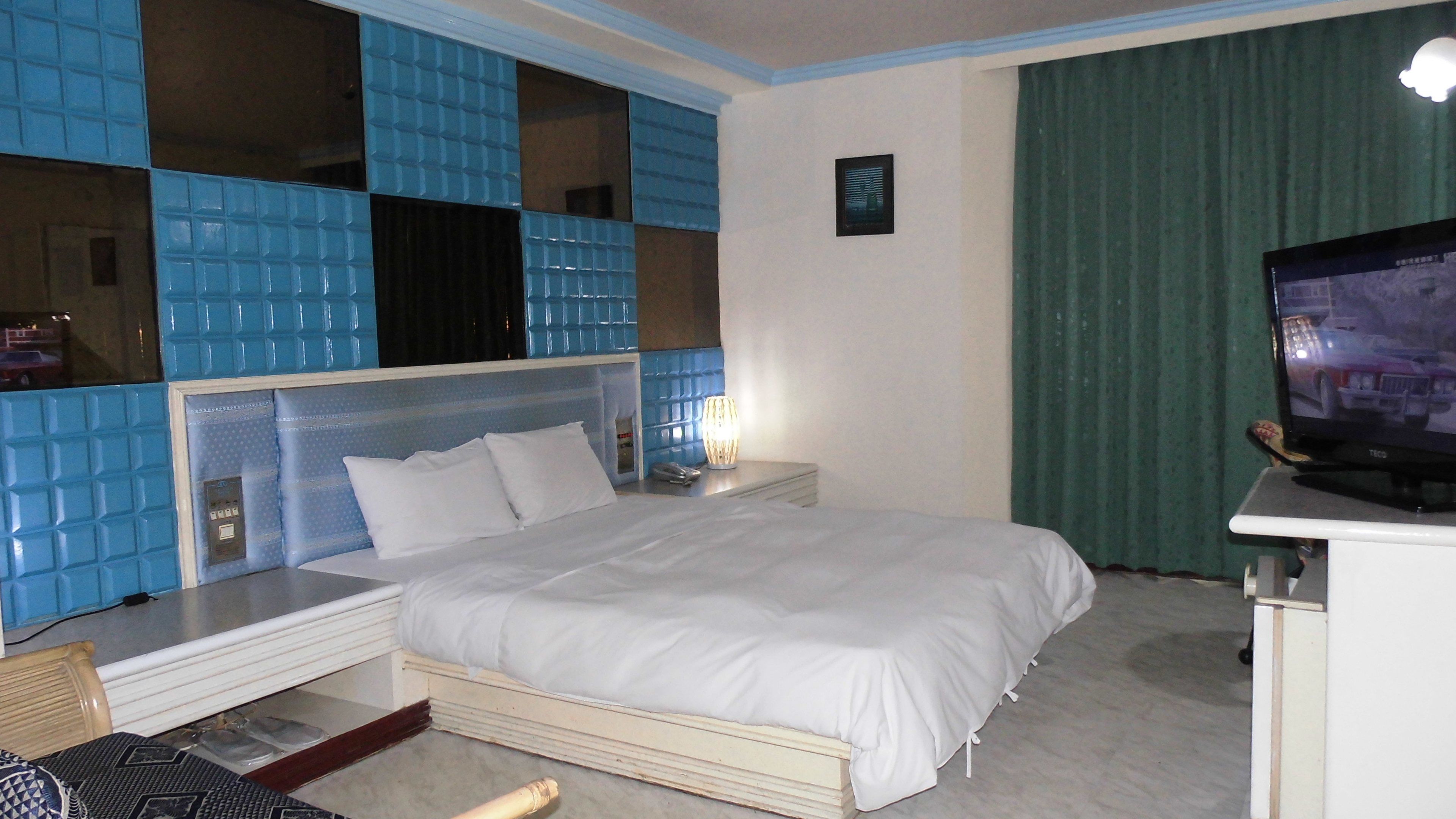 Ein Hotelzimmer mit Bett, Nachttisch, Fernseher und Fenster mit blau gefliesten Wänden.