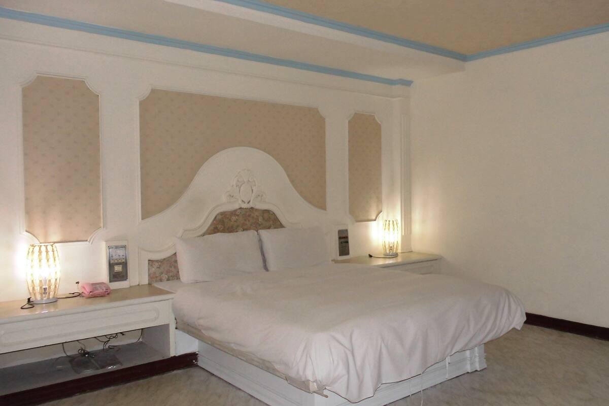 Una habitación de hotel con una cama grande, dos mesitas de noche, un abajur y un termostato en la pared.