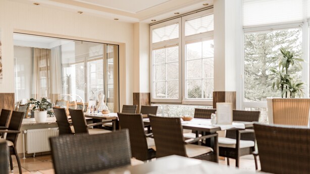 Un comedor con mesas y sillas, ventanas amplias y vistas a árboles fuera.
