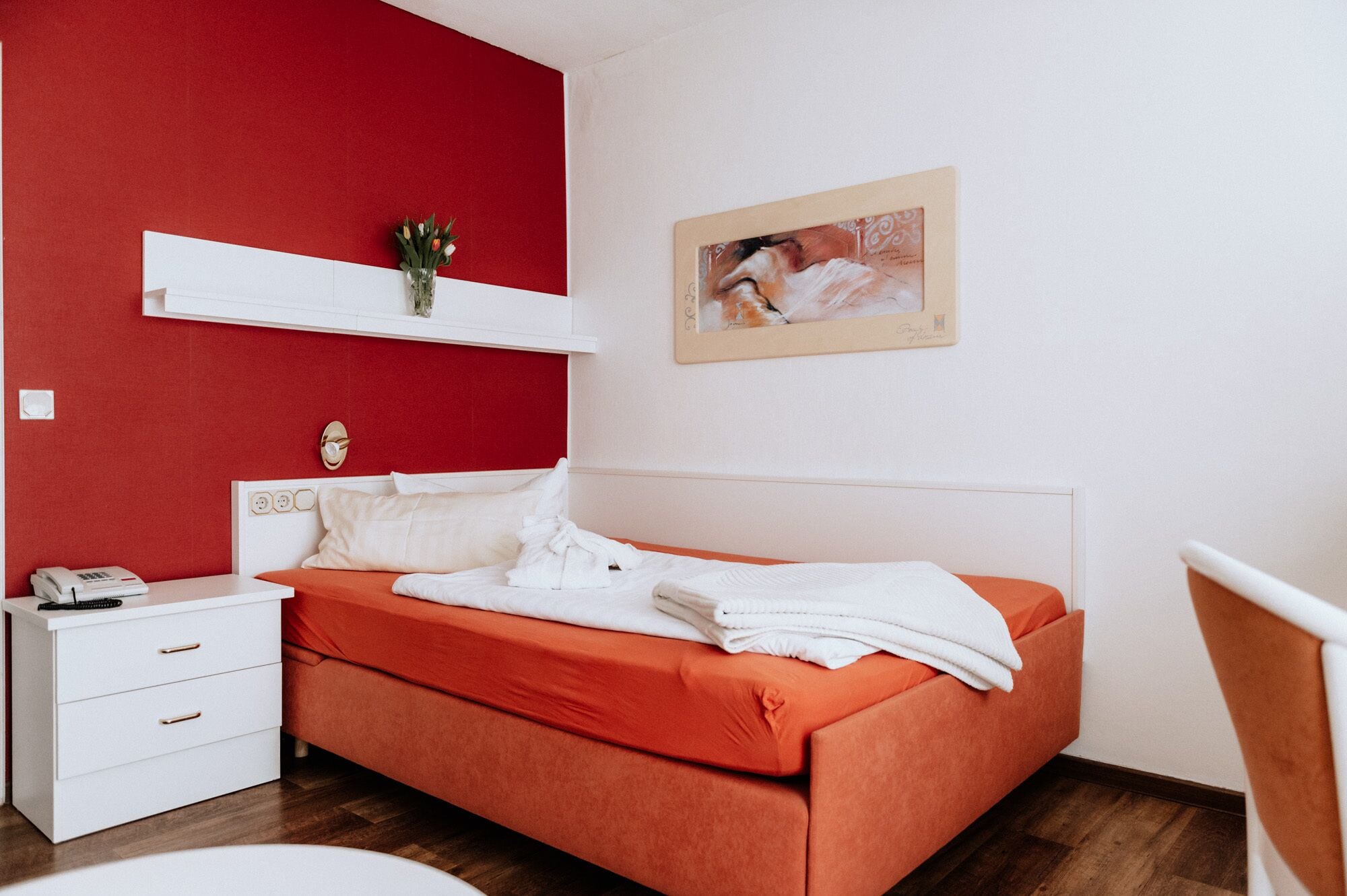 Un dormitorio con una pared de acento roja, una mesita de noche blanca con un teléfono, una cama con ropa de cama naranja y un cuadro en la pared.