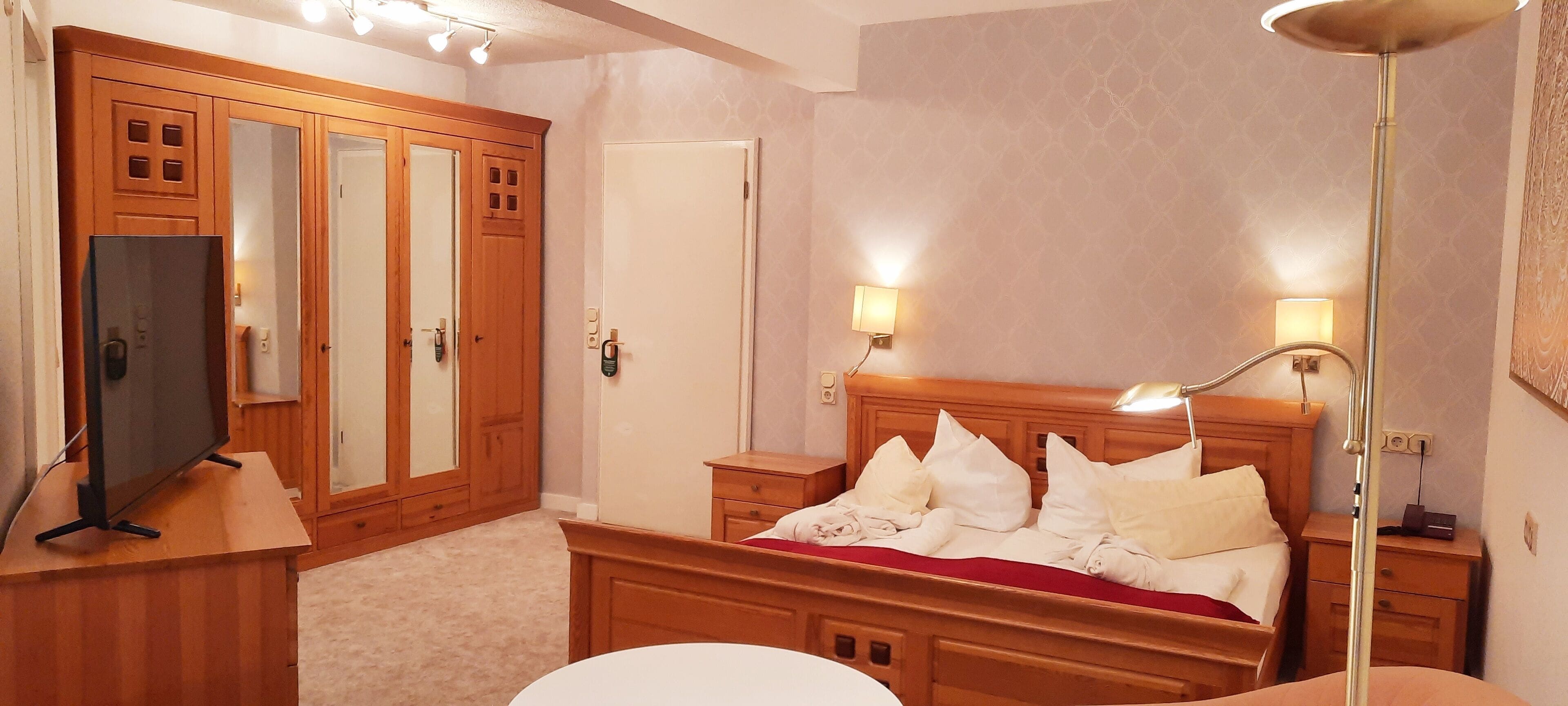 Een hotelkamer met een houten garderobe, een televisie, een bed met wit beddengoed en nachtlampjes.
