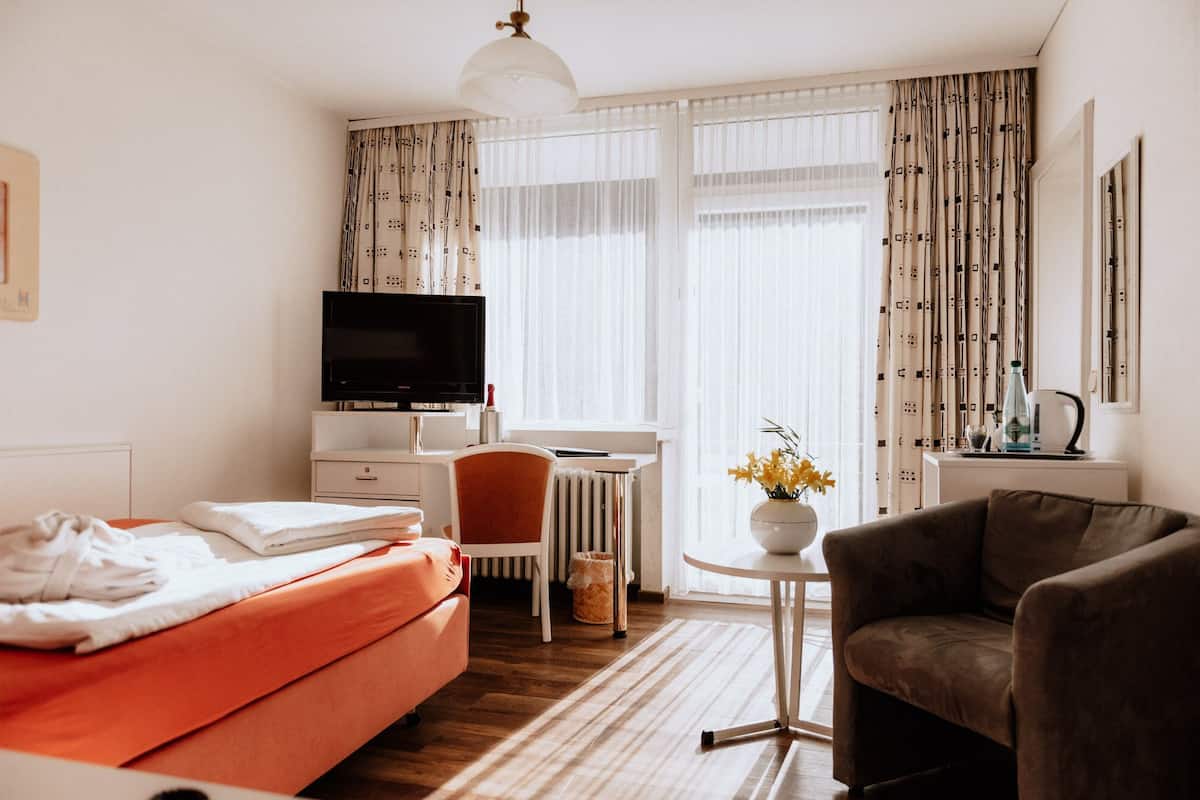 A hotel room with a bed, a desk with a television, a chair, a small table with a vase of flowers, and a radiator.