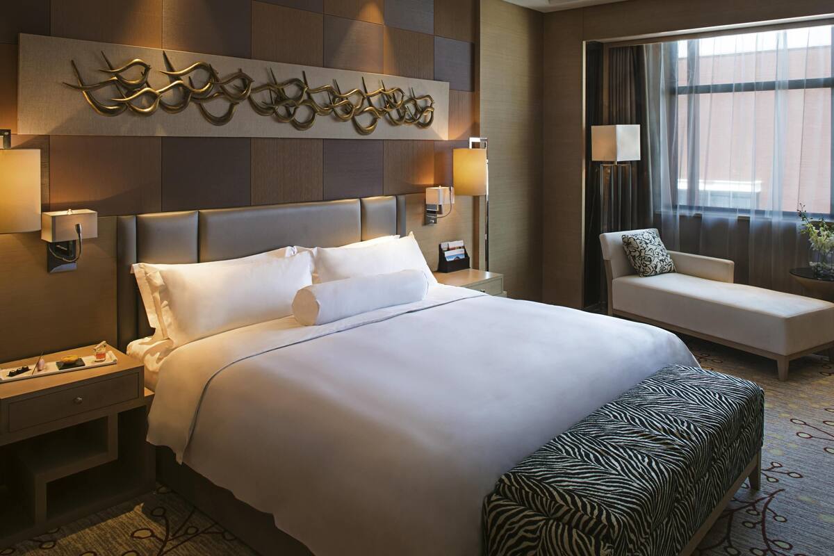 Una habitación de hotel con una cama grande, un sofá, un reposapiés estampado con motivos de cebra, una obra de arte en la pared y una ventana con cortinas.