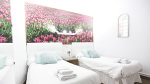 A hotel room with two beds, white bedding, a large floral mural on the wall, a small table with a vase, and a window.