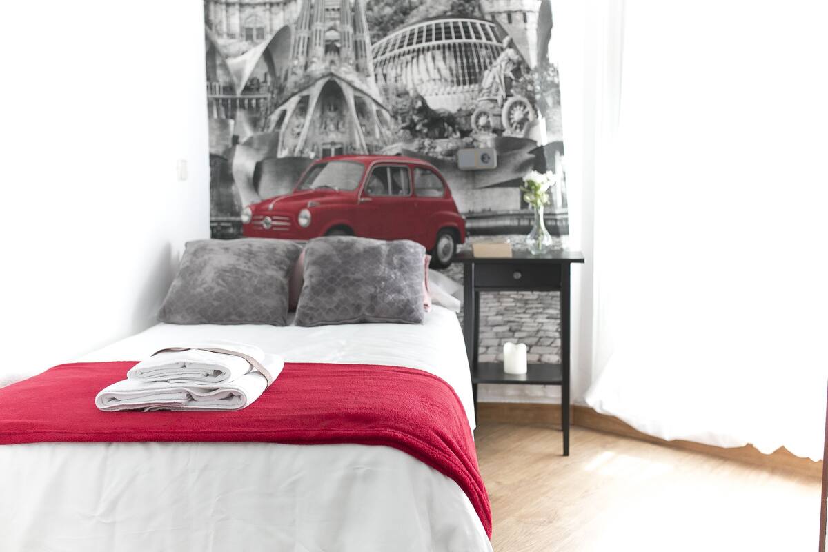 Una cama bien hecha con un edredón rojo y toallas dobladas, una mesita con un florero, y un mural en la pared que muestra un coche antiguo y detalles arquitectónicos.