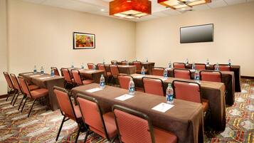 A conference room with multiple tables and chairs, water bottles on each table, a flat-screen TV on the wall, and a patterned carpet.
