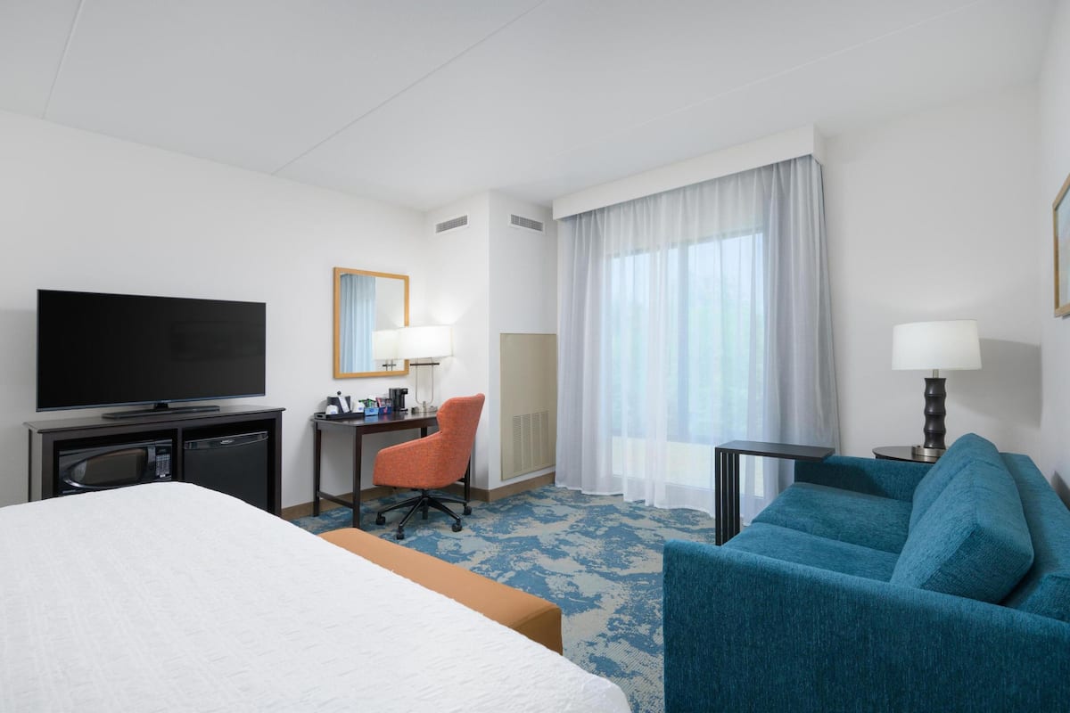 A hotel room with a bed, a desk with a computer, a TV, a sofa, and a window with curtains.
