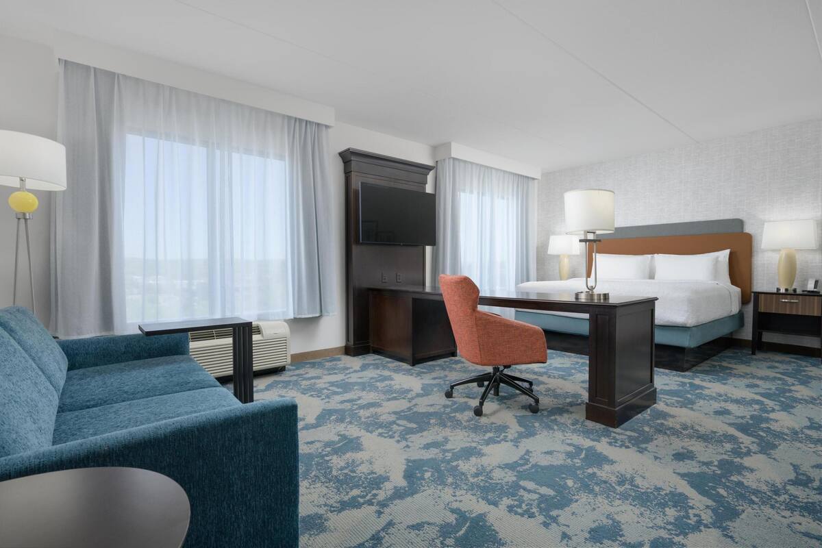 A hotel room with a bed, a desk, a chair, a TV, and a lamp.