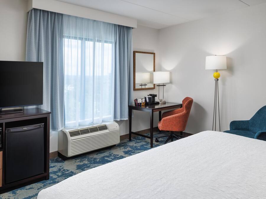 A hotel room with a bed, a desk, a chair, a television, a lamp, and a heater.