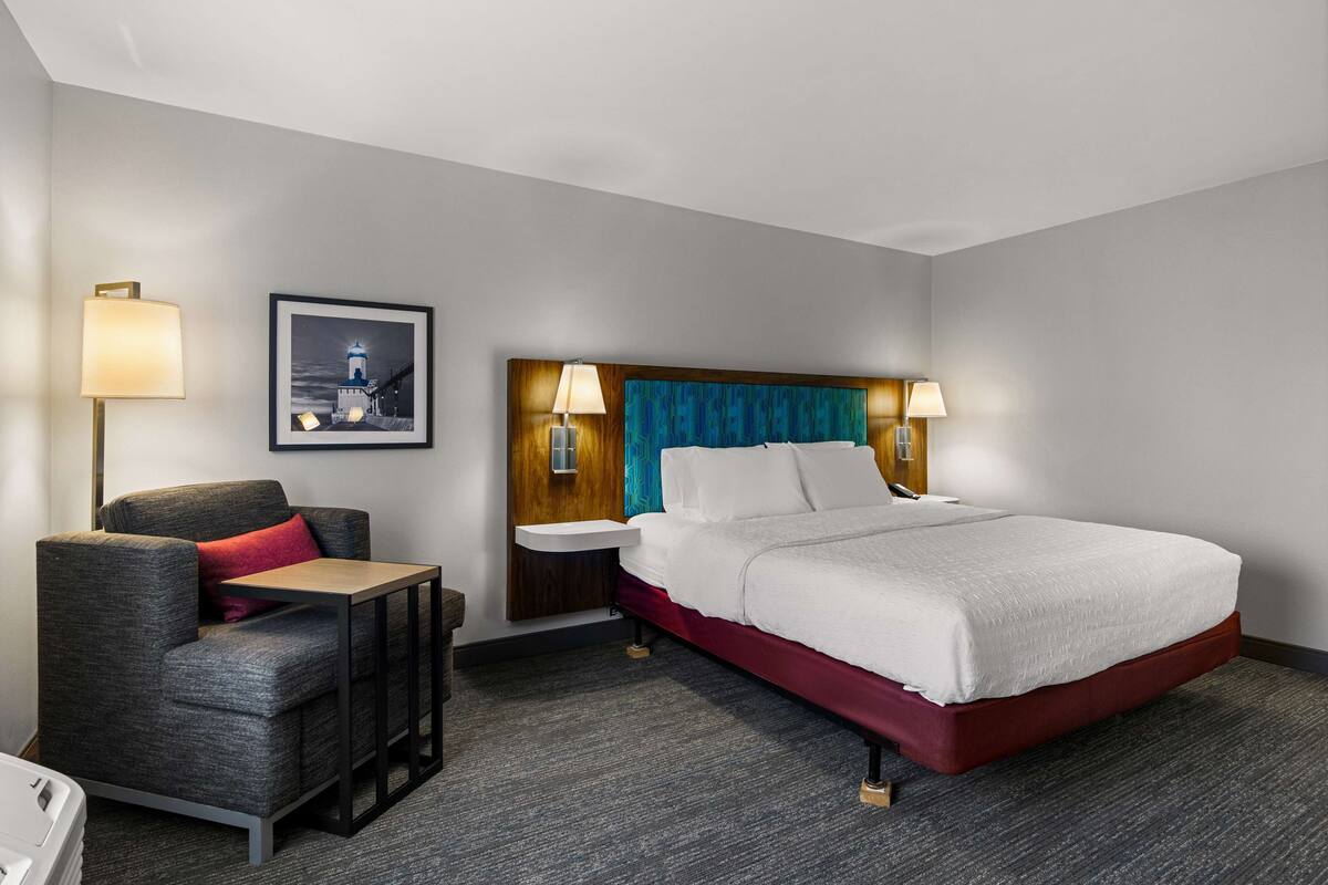 A hotel room with a bed, a chair, a small table, and a nightstand with lamps.