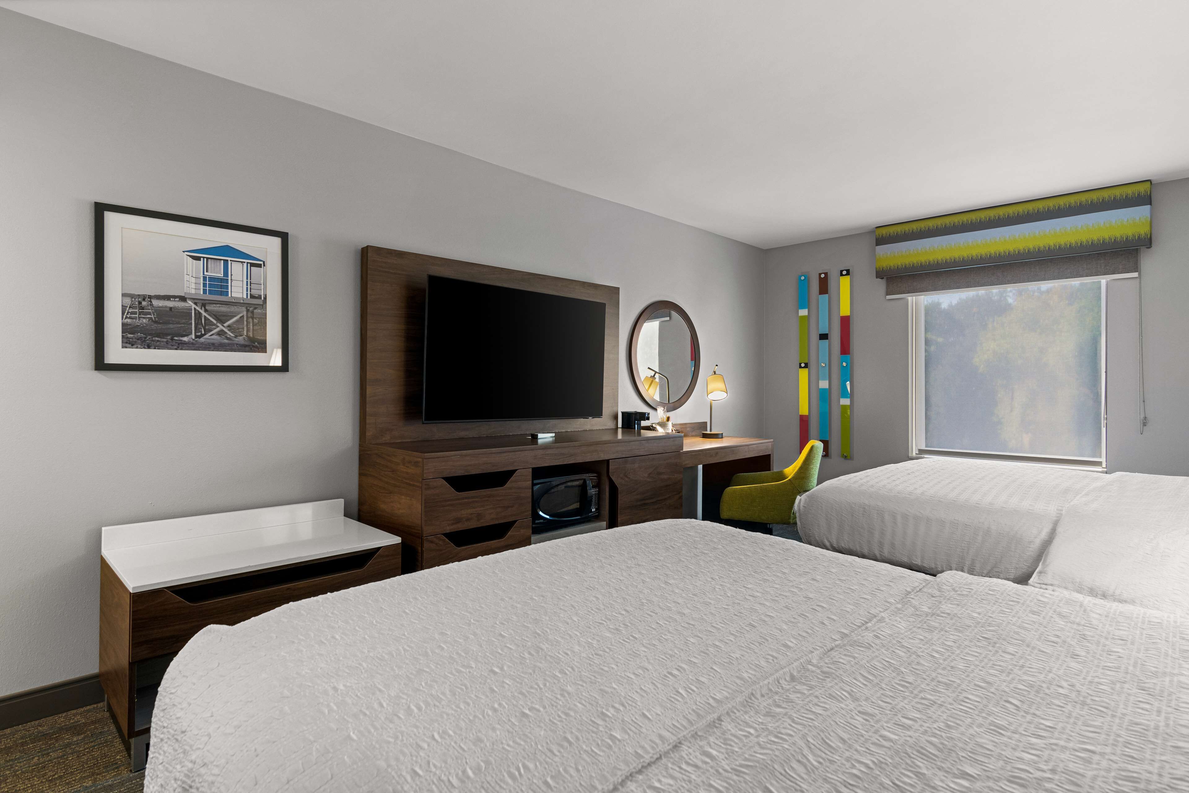A hotel room with two beds, a TV, a desk with a mirror, a framed picture on the wall, and a window with a view.
