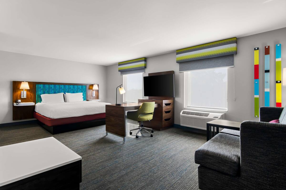 A hotel room with a bed, desk, chair, TV, and air conditioning unit.