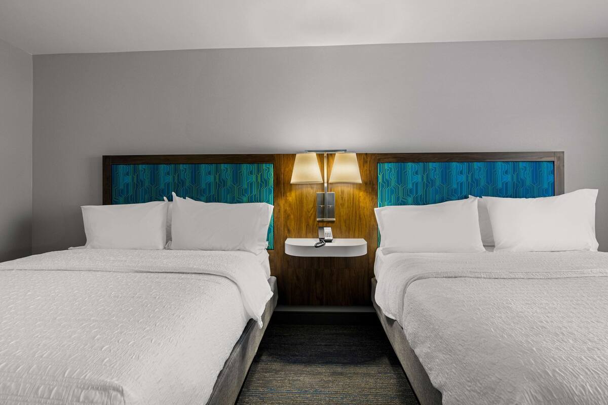 A hotel room with two beds, a nightstand with a lamp, and a wall-mounted sink.