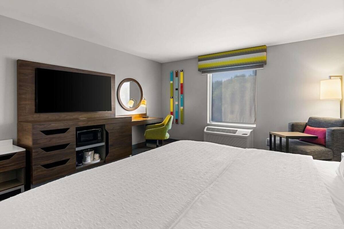 A hotel room with a large bed, a flat-screen TV, a desk with a chair, a window with a view, and a small table.