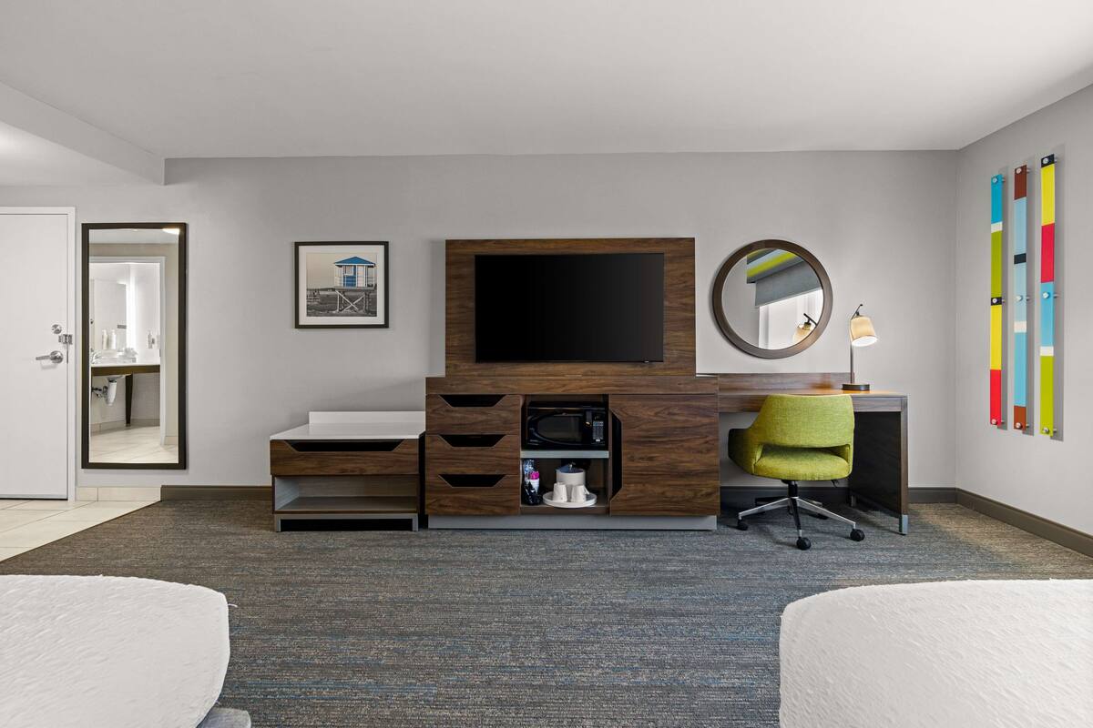 A hotel room with a bed, a desk with a TV, a microwave, a chair, and a mirror.