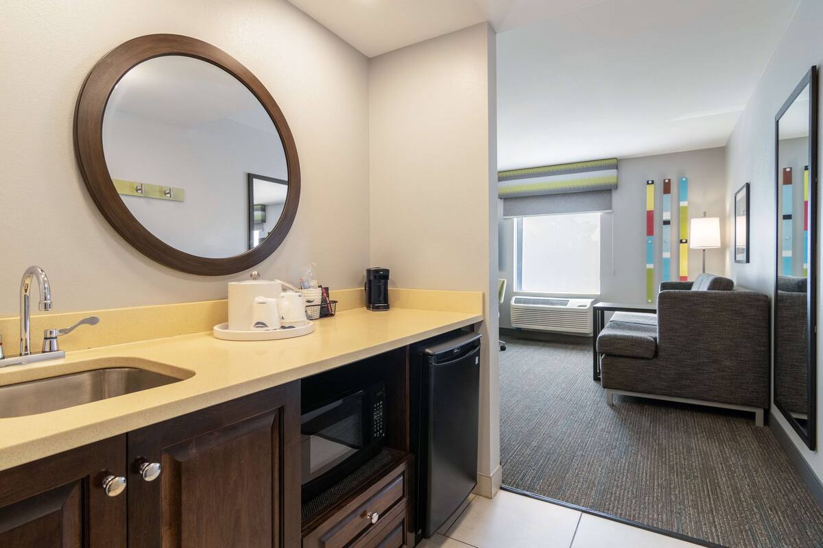 A modern hotel room with a kitchen area, including a sink, microwave, and coffee maker. There is a seating area with a couch and a view of the outdoors.