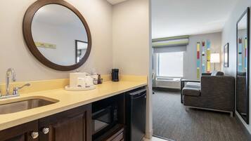 Habitación de hotel moderna con espacio para cocina, que incluye un lavamanos, microondas y cafetera. Hay una zona de estar con sofá y vistas al exterior.