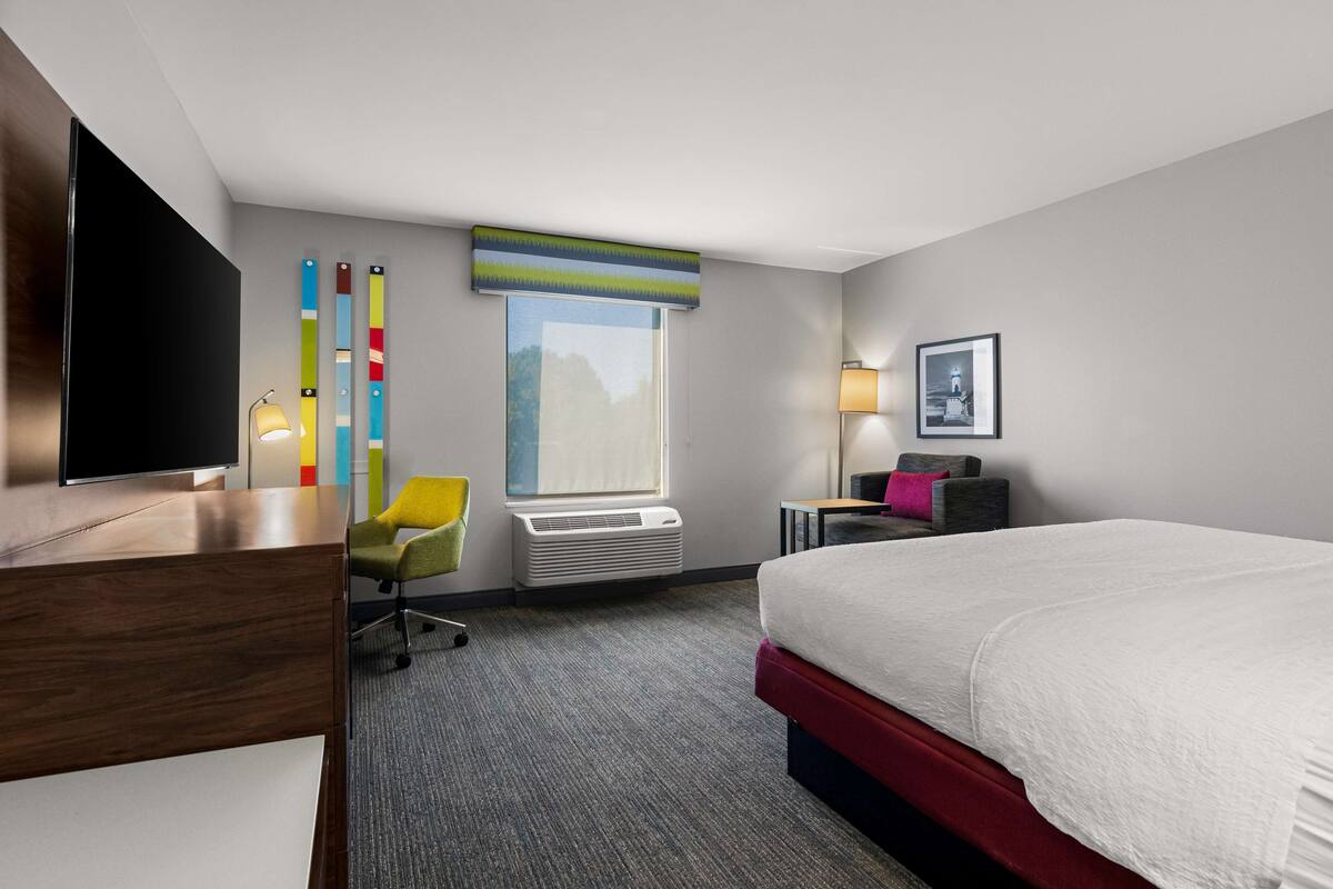 A hotel room with a bed, a desk, a chair, a window, and a wall-mounted TV.