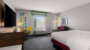 Una habitación de hotel con cama, escritorio, silla, ventana y televisor instalado en la pared.