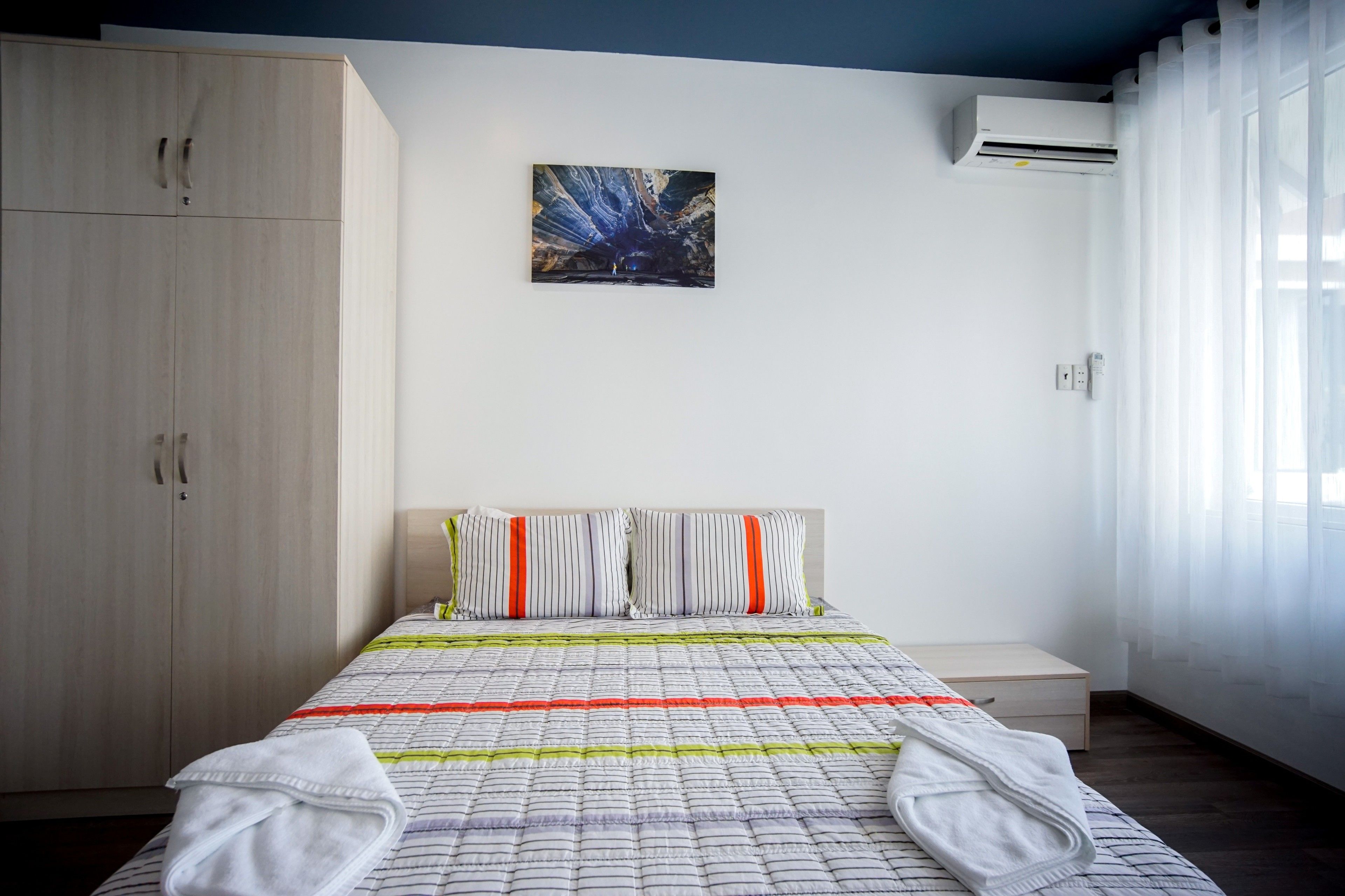 A bedroom with a bed, a wardrobe, a painting, and an air conditioning unit.