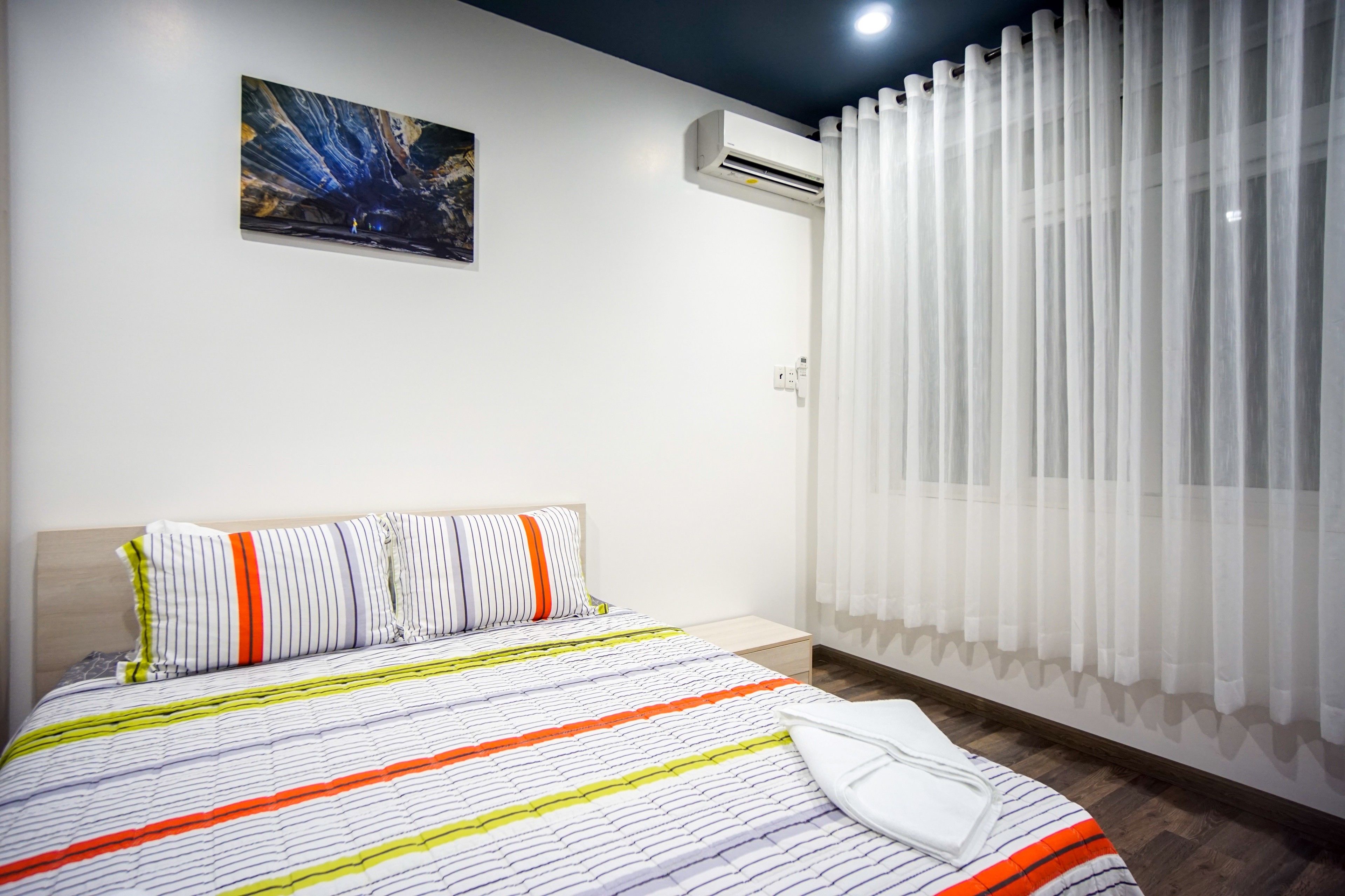 A hotel room with a bed, a painting on the wall, an air conditioning unit, and sheer curtains.