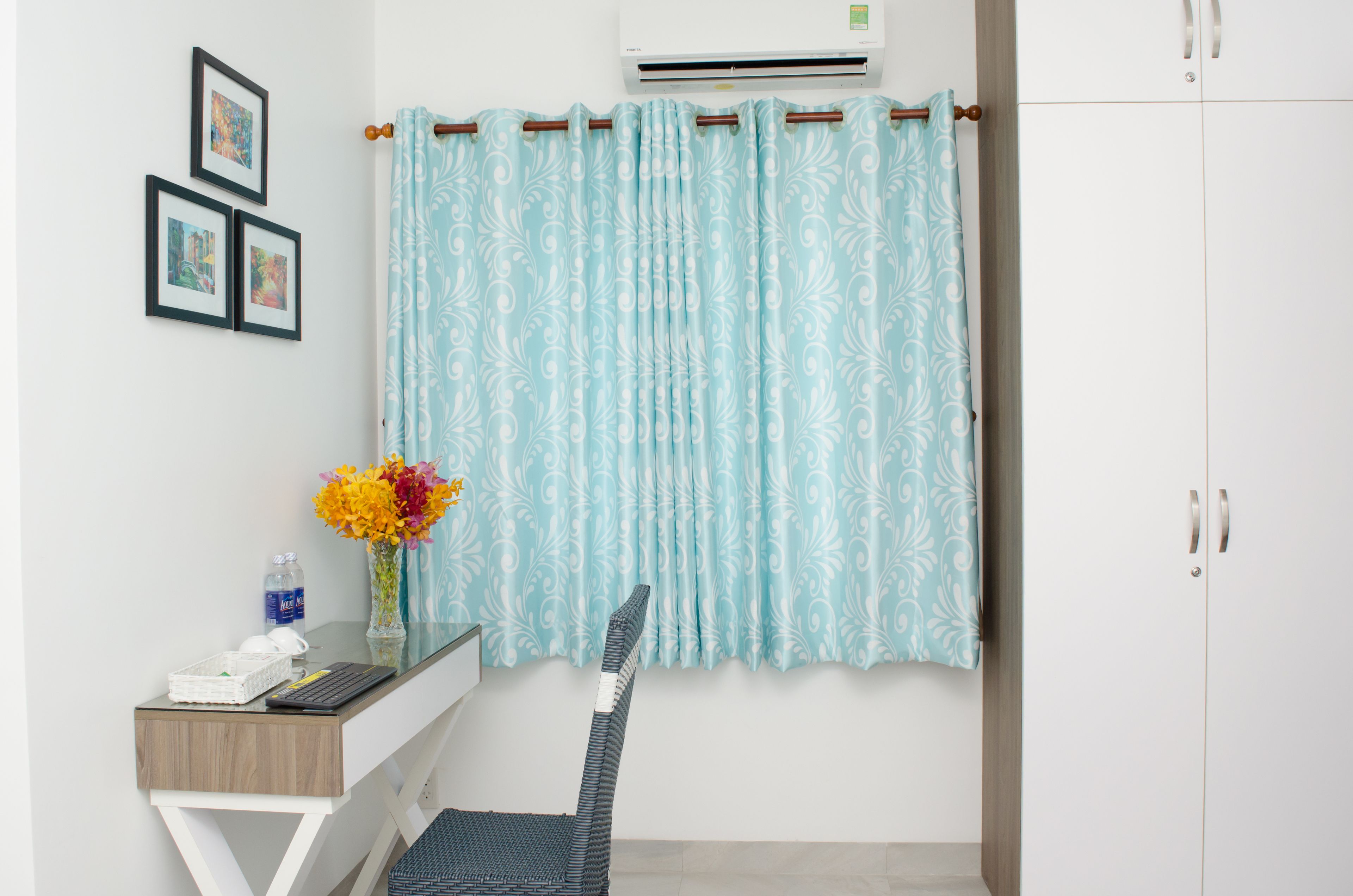 A room with a white wardrobe, a desk with a chair, a small table with a vase of flowers, and a curtain with a floral pattern.