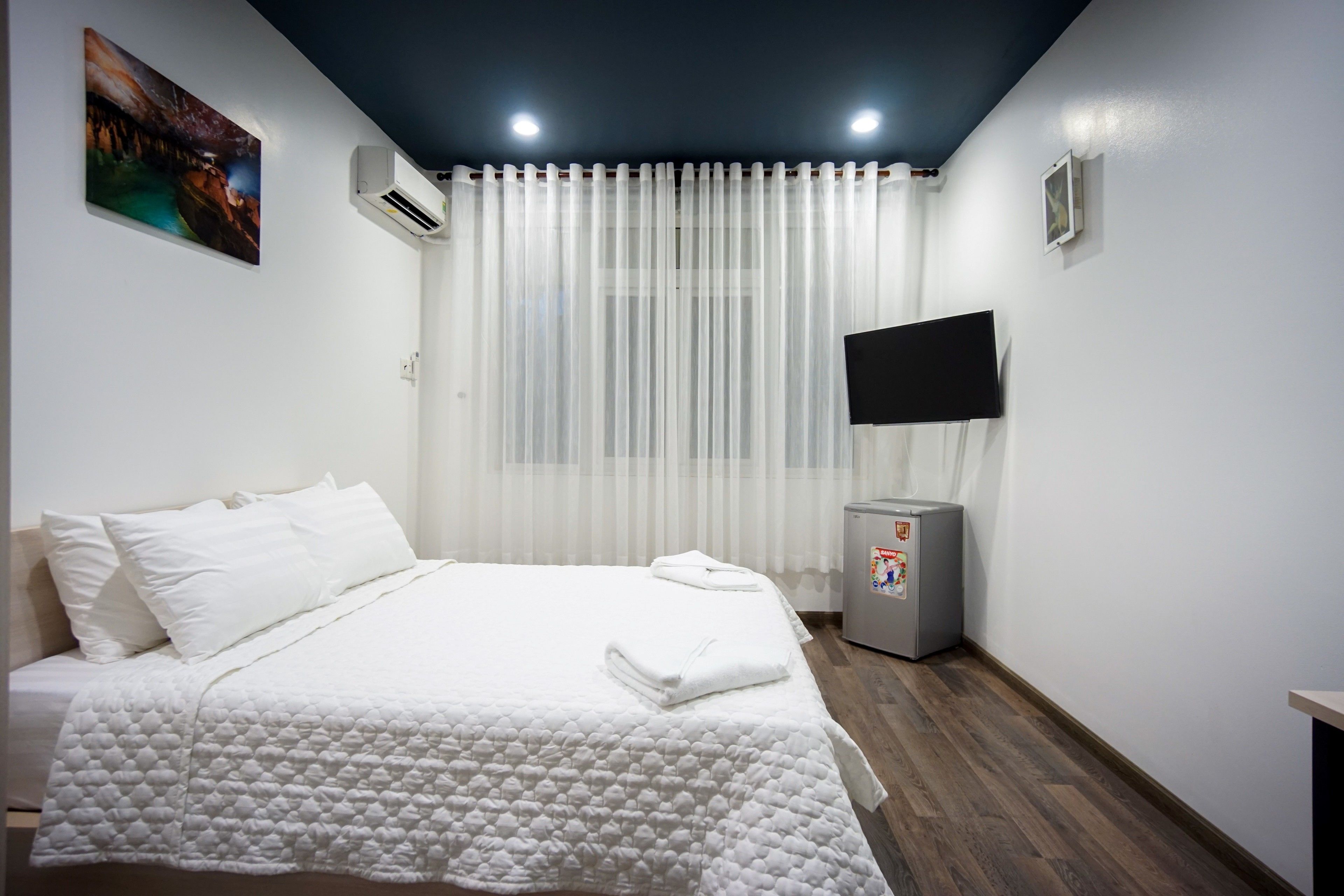 A hotel room with a bed, white curtains, a flat-screen TV, and a refrigerator.