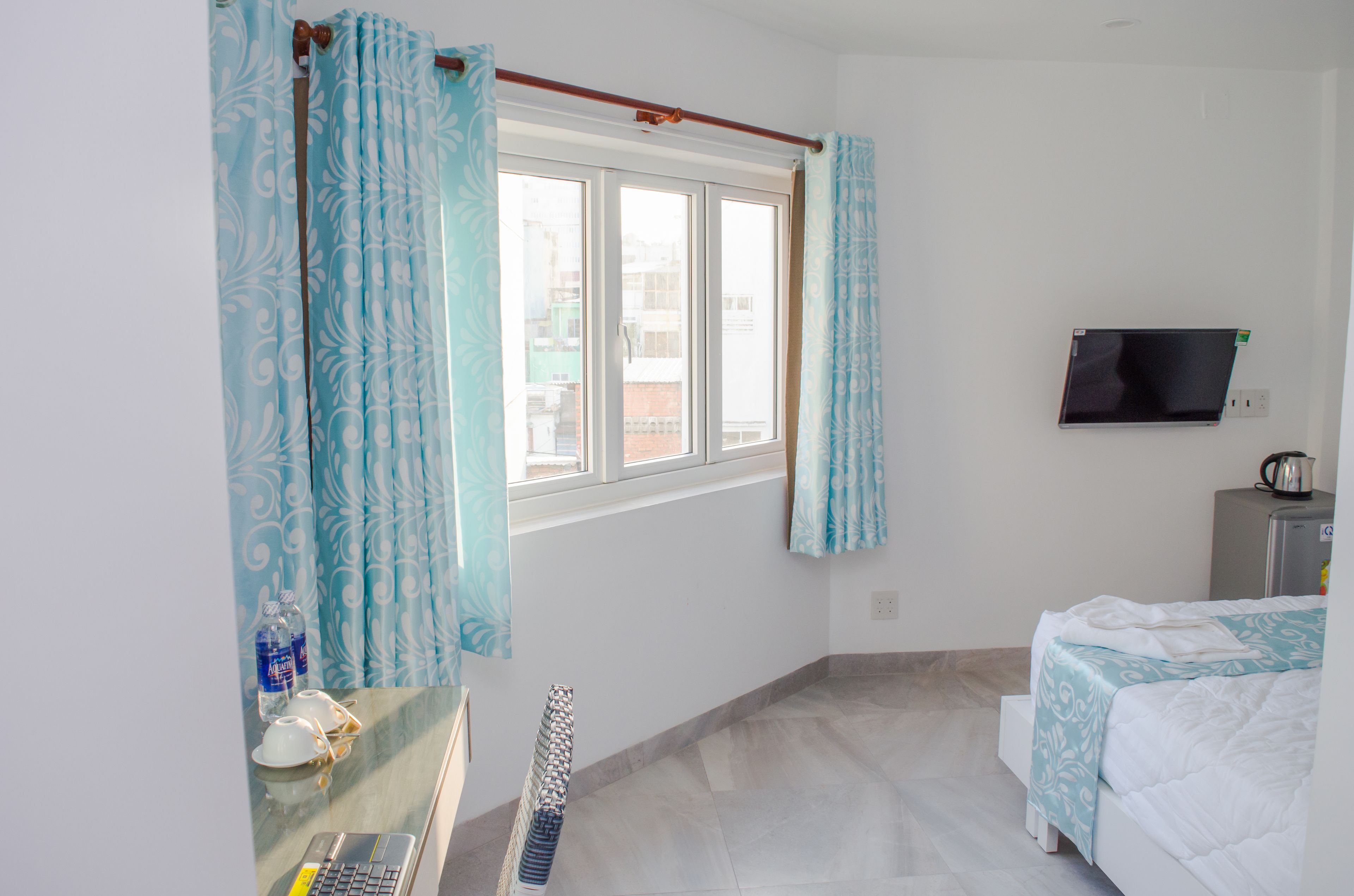 Habitación de hotel con cama, televisión montada en la pared, ventana con cortinas, radiador y una mesa con una botella y vasos.