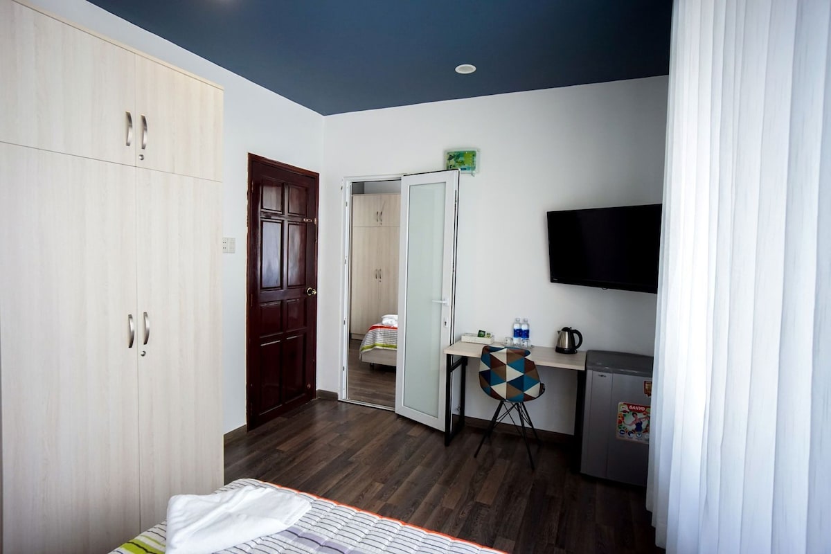 A bedroom with a bed, a wardrobe, a desk with a chair, a television, and a bathroom visible through an open door.