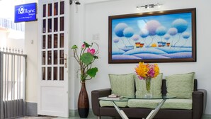 A modern living room with a glass coffee table, a brown sofa with green cushions, a large framed painting, and a potted plant. - LeBlanc Saigon (Ho Chi Minh City)