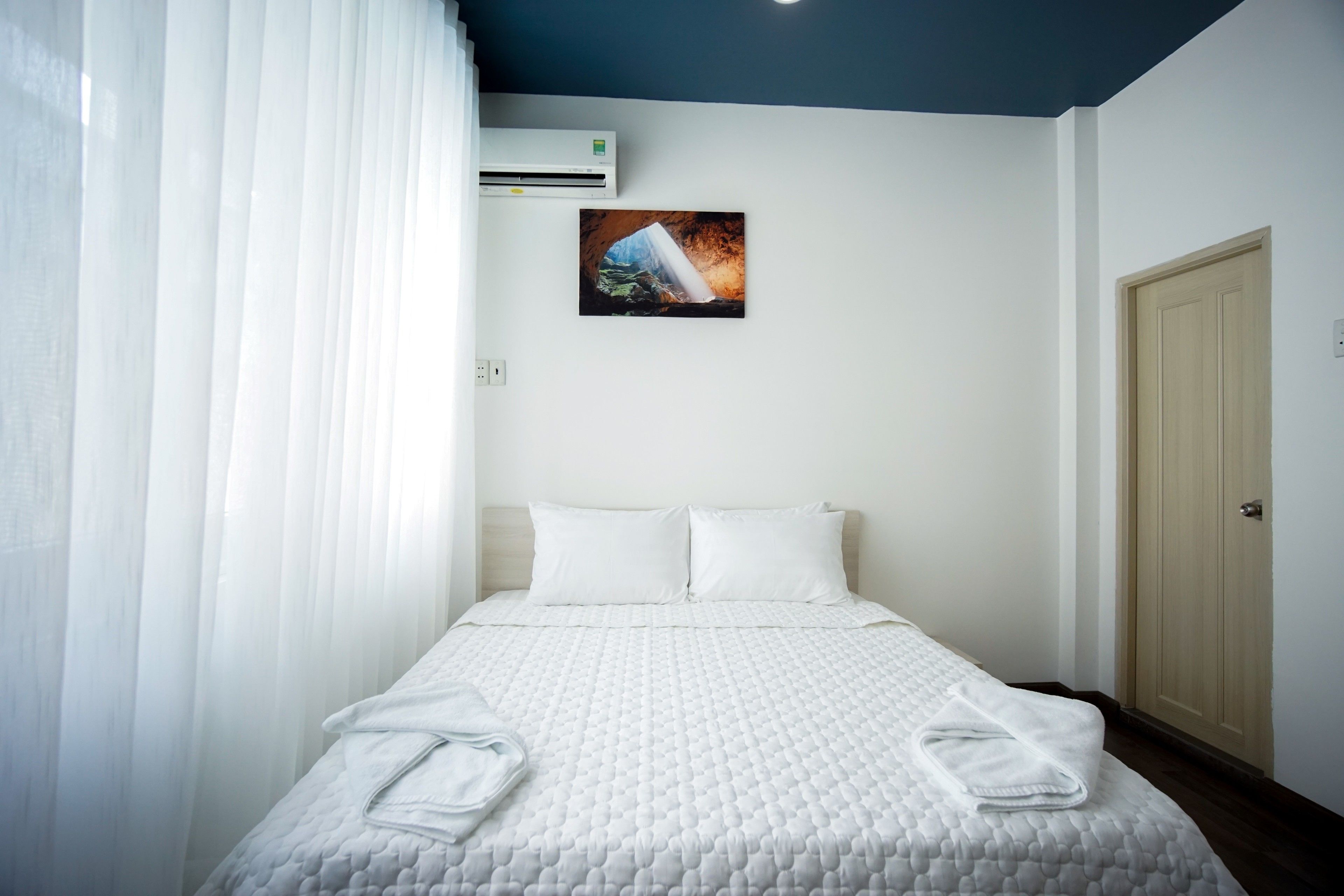 A hotel room with a bed, white linens, a painting on the wall, and an air conditioning unit.