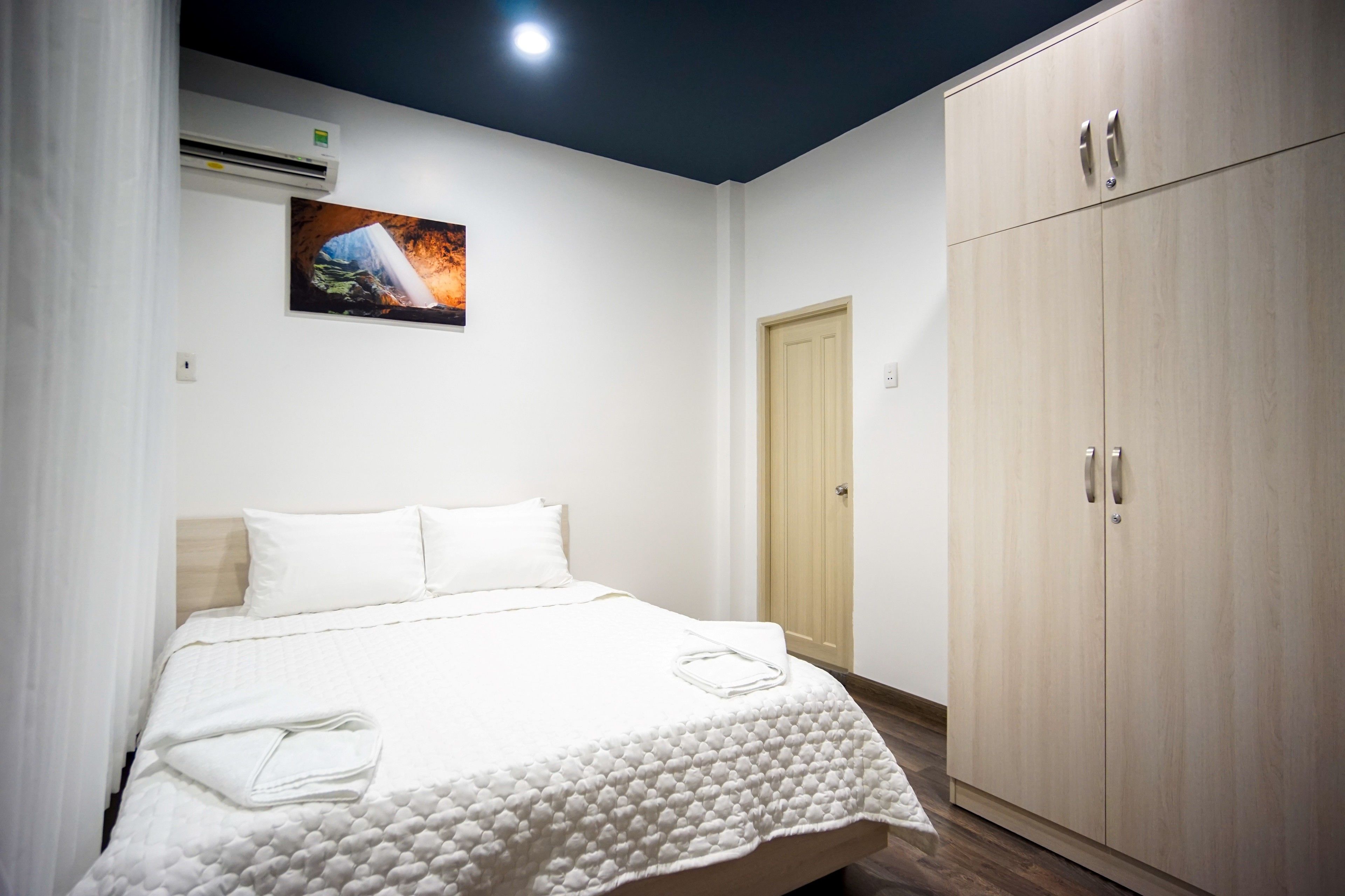 A neatly made bed with a white duvet and pillows, a wooden wardrobe with two doors, and a framed picture on the wall.