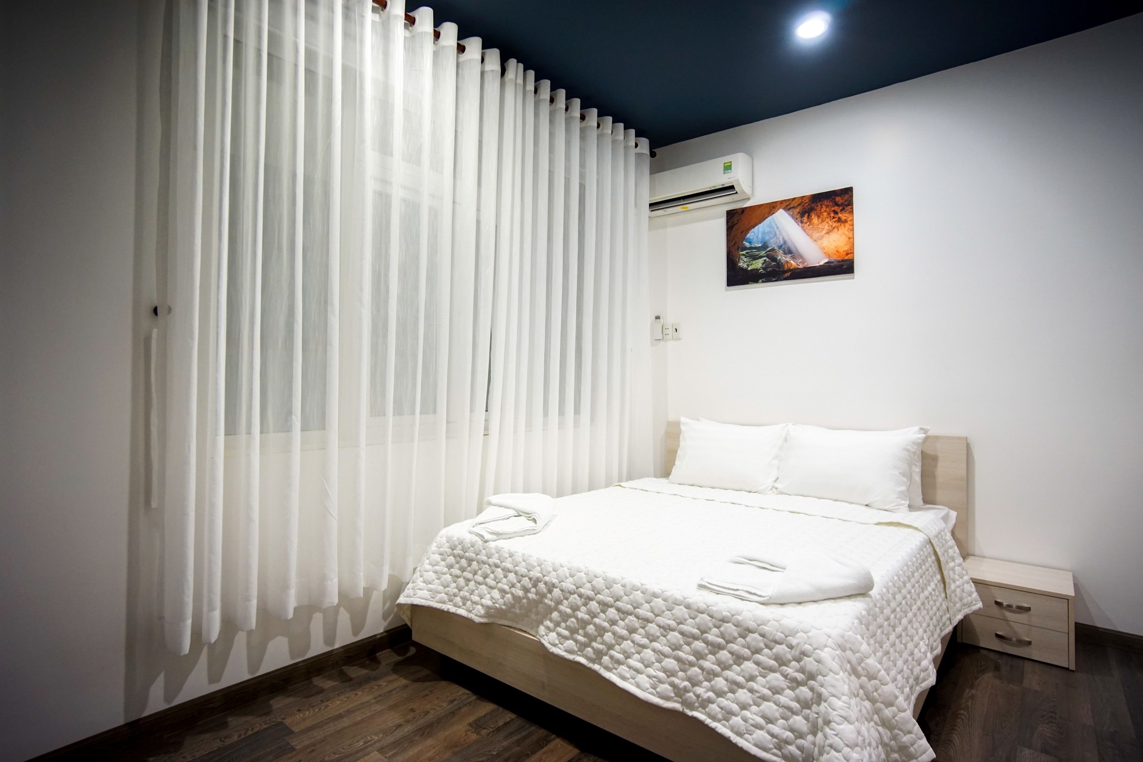 Een net opgemaakt bed met een witte deken en kussens, een nachtkastje met een lamp, en een wandairconditioner.
