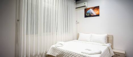 A neatly made bed with a white duvet and pillows, a bedside table with a lamp, and a wall-mounted air conditioner.