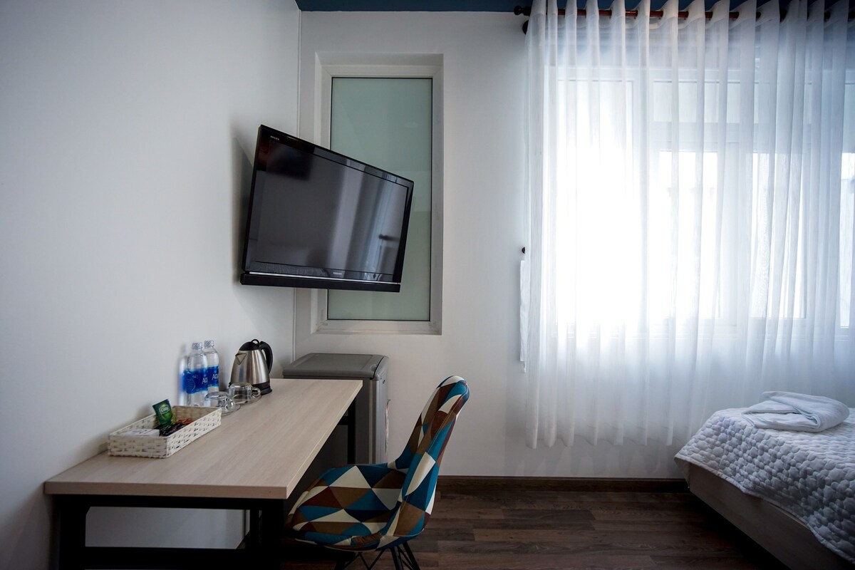 A room with a bed, a TV mounted on the wall, a desk with a chair, and a window with sheer curtains.