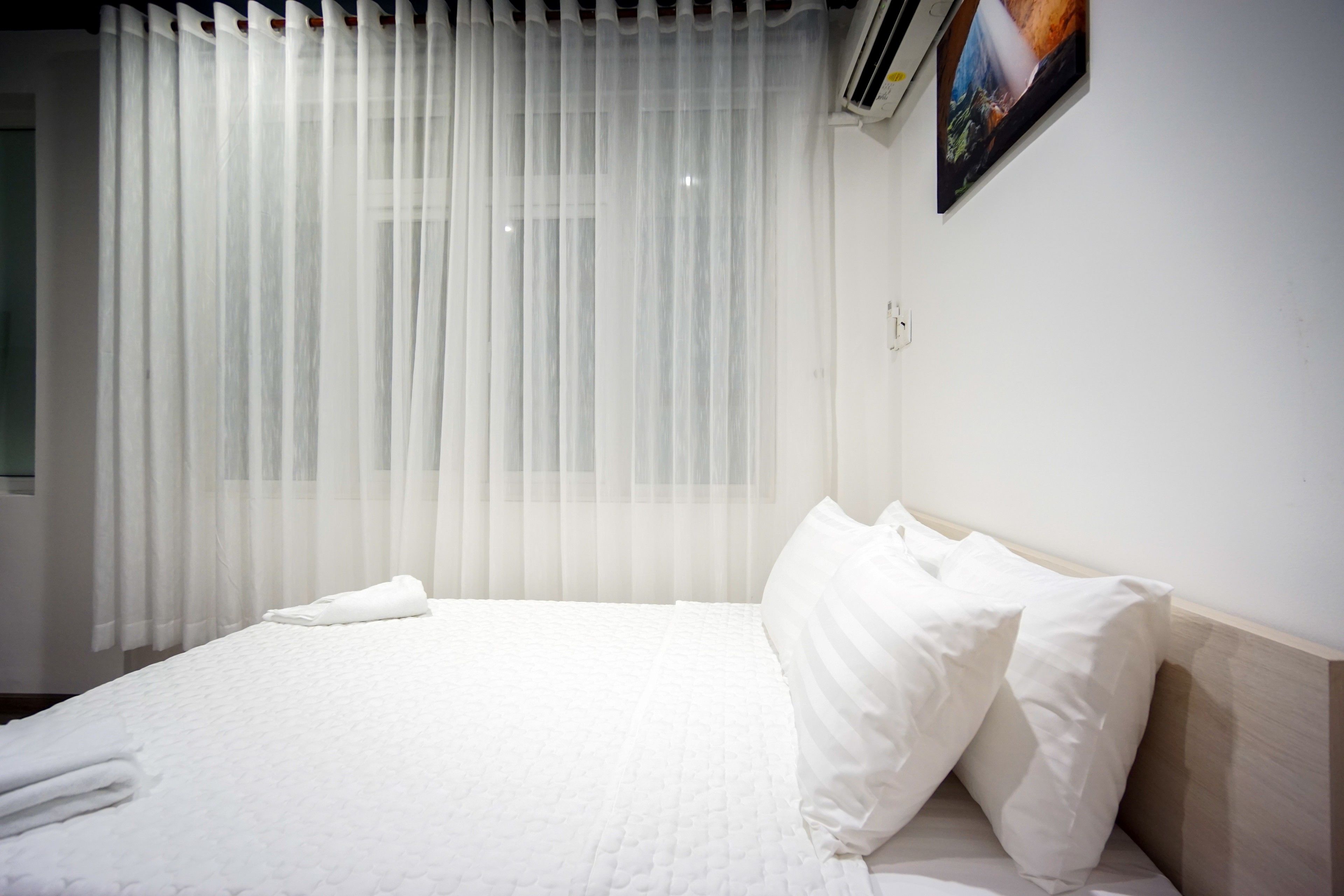 A neatly made bed with white linens and pillows, a white wall, and a window with sheer curtains.