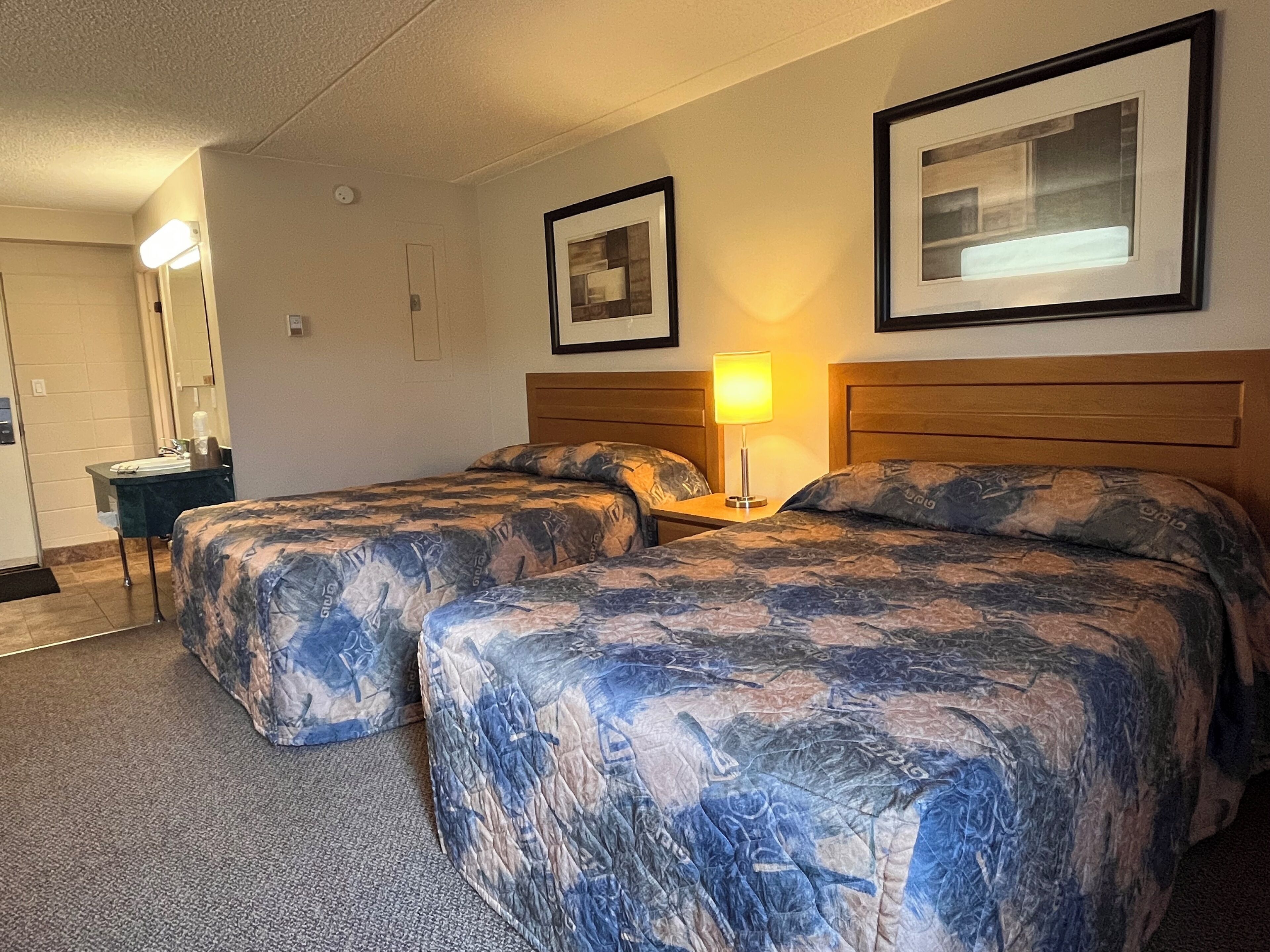 Habitación de hotel con dos camas, una mesita de noche con lámpara, un espejo y cuadros en la pared.