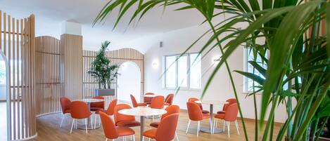 A modern interior with round tables and orange chairs, wooden partitions, and large windows allowing natural light.