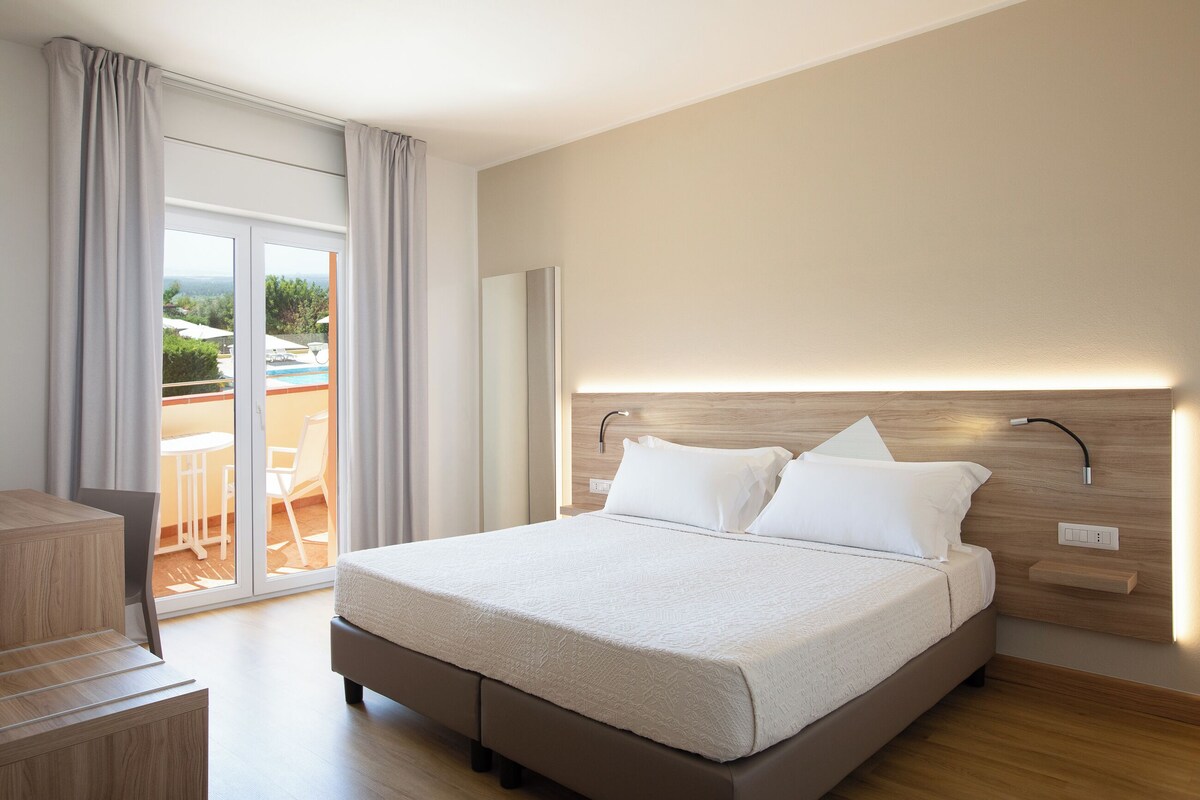Habitación de hotel moderna con una cama grande, un cabecero de madera y vistas a una piscina y un área de estar exterior a través de una puerta corrediza de vidrio.