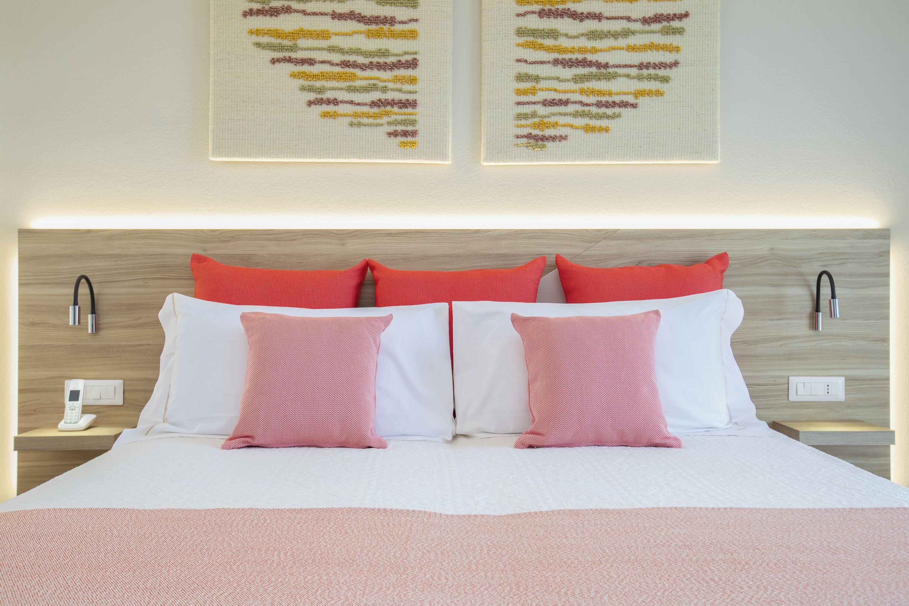 A neatly made bed with white and pink bedding, two wall-mounted bedside lamps, and two framed artworks above the bed.