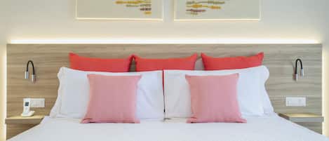 Ein ordentlich bezogenes Bett mit weißer und rosa Bettwäsche, zwei an der Wand befestigte Nachttischlampen und zwei gerahmte Kunstwerke darüber.