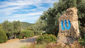 Een groot rotsbord waarop 'PORTOPINO HOTEL' staat, omringd door begroeiing en een weg.
