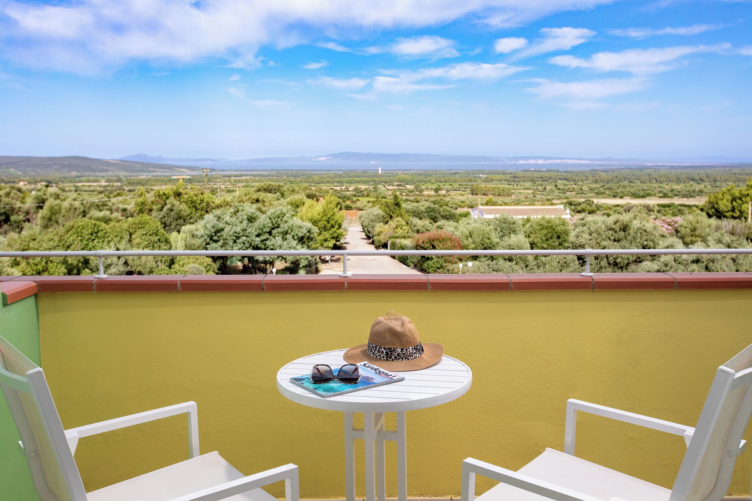 Un balcon avec une table, deux chaises blanches, un chapeau et des lunettes de soleil posés sur la table.