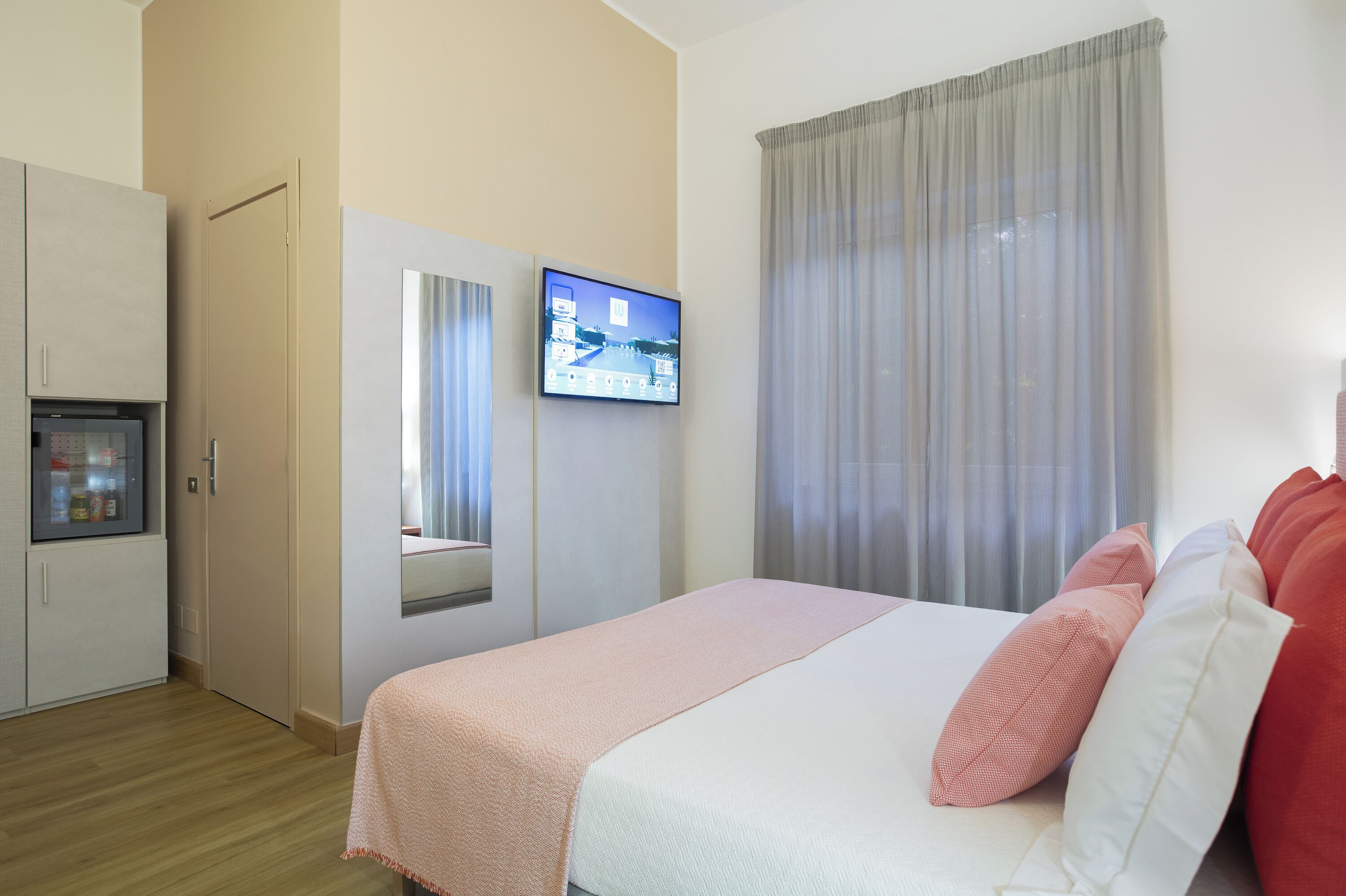 A hotel room with a bed, a television mounted on the wall, a wardrobe, and a window with curtains.